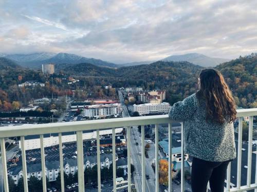 A person with curly hair stands on a balcony, overlooking a scenic mountain view with autumn foliage.