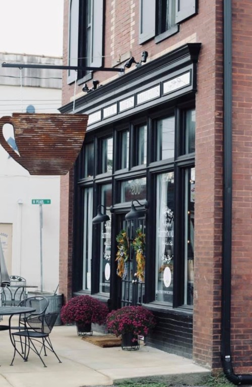 A charming storefront with large windows, a hanging cup sign, and flower pots outside, set in a brick building.