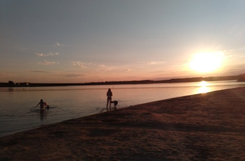 Silhouettes of people by a calm lake at sunset, with warm colors reflecting on the water and a sandy shore.