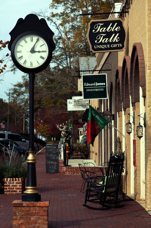 A street view featuring a clock, shops with signs, and a rocking chair on a brick sidewalk.