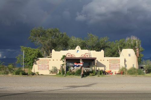 A southwestern-style building with a colorful sign, surrounded by trees, under a stormy sky with a hint of a rainbow.