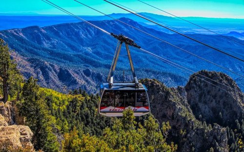 A cable car glides over a mountainous landscape, surrounded by lush greenery and distant peaks under a clear blue sky.