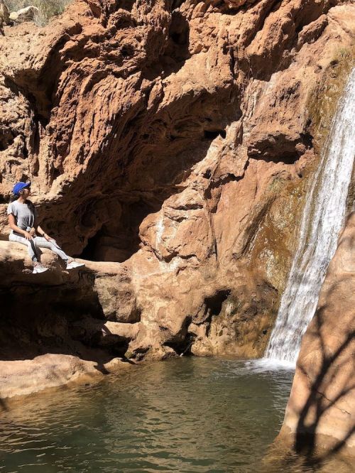 A person sits by a rocky waterfall, surrounded by natural scenery and a calm pool of water.