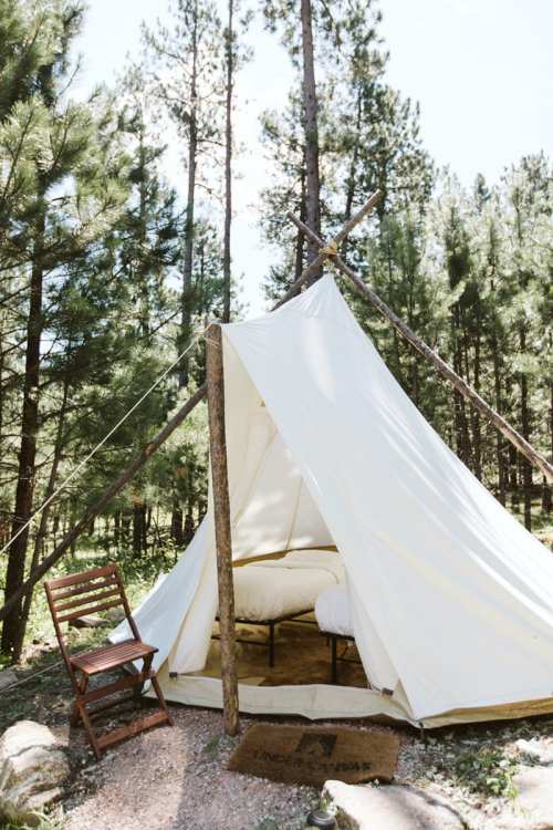 A white tent set in a forest, with a wooden chair nearby and trees surrounding the area.