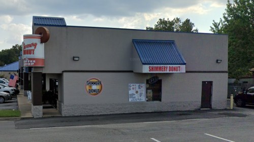 Exterior of Shimmery Donut shop with a blue roof, signage, and an open flag. Parking lot visible in front.