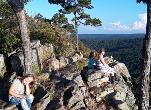 Three children sit on rocky terrain overlooking a lush green valley and blue sky, enjoying a scenic view.