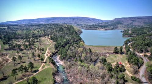 Aerial view of a serene landscape featuring a lake, lush greenery, and rolling hills under a clear blue sky.