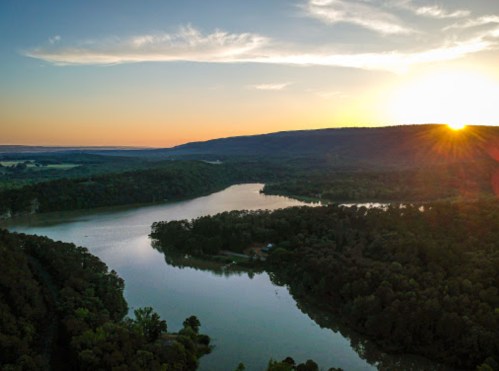 Aerial view of a serene lake surrounded by lush greenery at sunset, with mountains in the background.