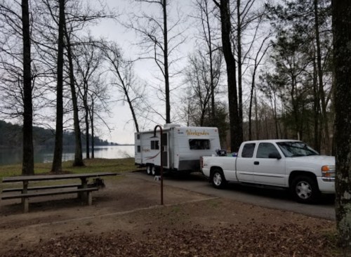 A white pickup truck towing a camper parked by a lake, surrounded by bare trees and a picnic table nearby.