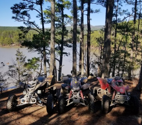 Three ATVs parked among trees by a lake, with a scenic view of hills in the background.