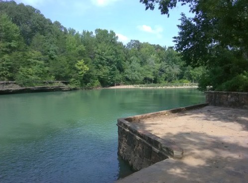 A serene river scene surrounded by lush green trees and a stone platform along the water's edge.