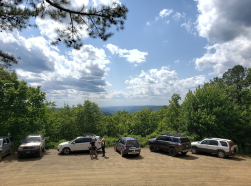 A scenic overlook with several parked cars, surrounded by lush greenery and a cloudy blue sky.