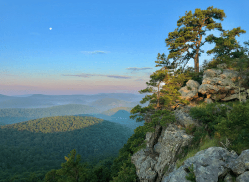 A scenic view of mountains and valleys under a clear blue sky, with a tree on a rocky ledge and a visible moon.