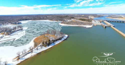 Aerial view of a river with ice and snow on one side, and clear water on the other, surrounded by trees and hills.