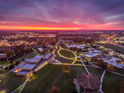 Aerial view of a campus at sunset, with vibrant colors in the sky and illuminated pathways winding through green spaces.