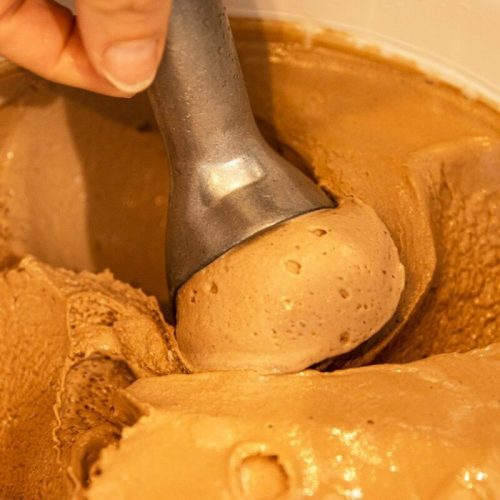 A hand holding an ice cream scoop, lifting a scoop of creamy chocolate ice cream from a container.