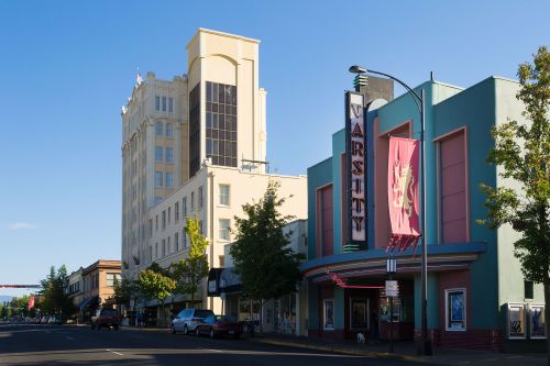 A colorful theater marquee for "Varsity" next to a tall building, with trees and parked cars lining the street.