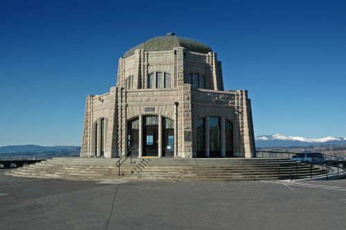 A stone building with a dome, set against a clear blue sky and snow-capped mountains in the background.