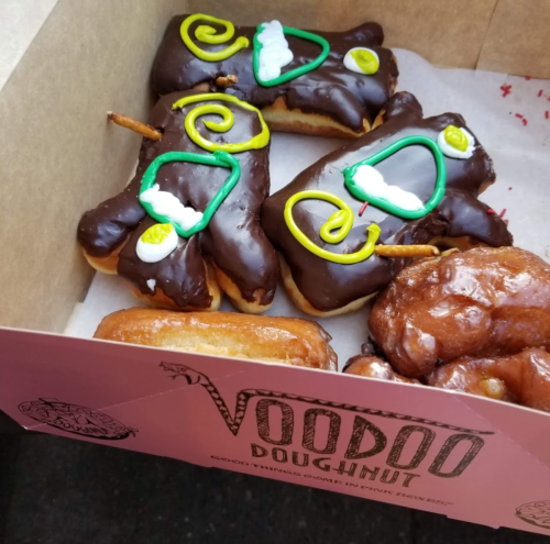A box of colorful doughnuts, including chocolate-covered ones decorated with green and white icing, resembling festive characters.