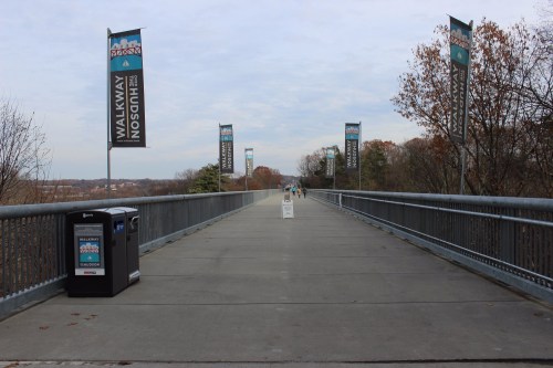 A wide walkway with banners on either side, leading into the distance, surrounded by trees and a trash bin nearby.