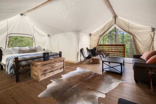 Cozy glamping tent interior with a bed, seating area, wooden furniture, and large windows overlooking nature.
