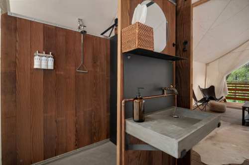 A rustic outdoor shower with wooden walls, a concrete sink, and toiletries on display in a tented space.