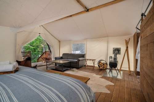 Cozy glamping tent interior with a bed, seating area, wood stove, and wooden accents in a natural setting.
