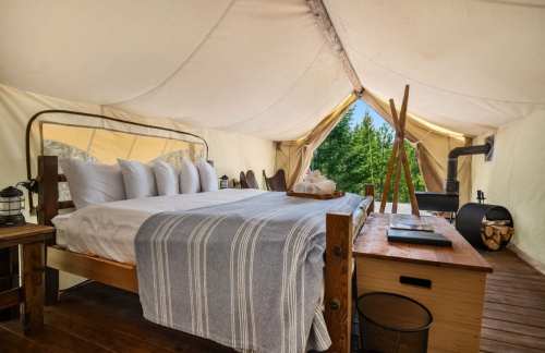 Cozy glamping tent interior with a bed, wooden furniture, and a view of trees outside.
