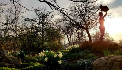 Silhouette of a statue holding a basket, surrounded by blooming flowers and trees at sunset.