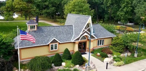 A charming building with a peaked roof, surrounded by greenery and a flag, set in a park-like environment.