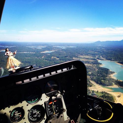 Aerial view from a helicopter cockpit, featuring a dashboard and a hula dancer figurine against a scenic landscape.