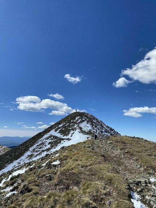 A snow-capped mountain peak under a clear blue sky with a few clouds, featuring a hiking trail leading to the summit.