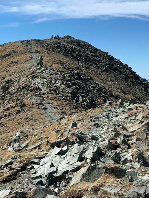 A hiker ascends a rocky mountain trail under a clear blue sky, with other hikers visible at the summit.