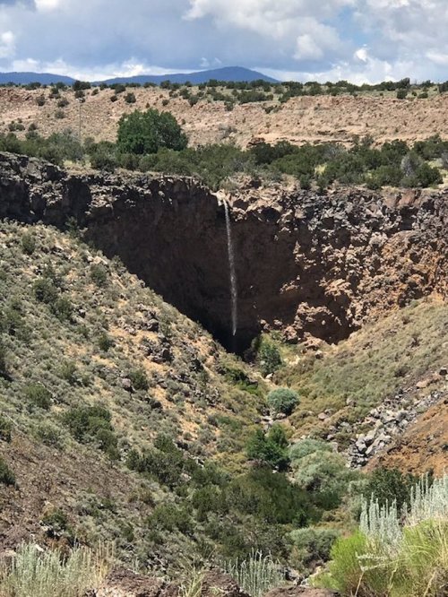A waterfall cascades into a rocky canyon surrounded by greenery and distant mountains under a partly cloudy sky.