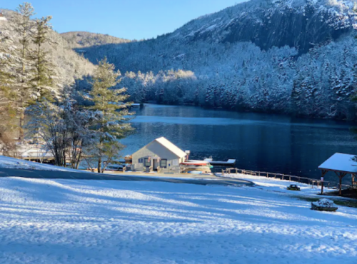 A serene winter scene featuring a snowy landscape, a calm lake, and a small cabin surrounded by trees and mountains.