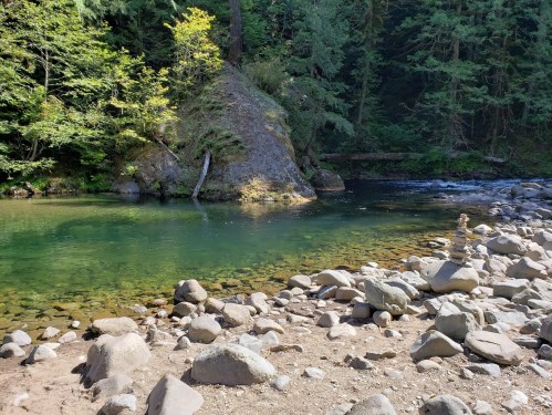 A serene river scene with clear water, rocky shore, and lush green trees in a peaceful natural setting.