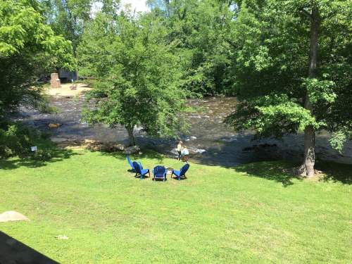 A serene riverside scene with green grass, trees, and blue chairs, featuring a person wading in the water.
