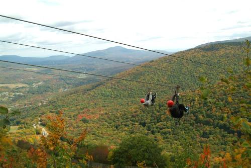 Two people ziplining over a vibrant, autumn-colored landscape with mountains in the background.