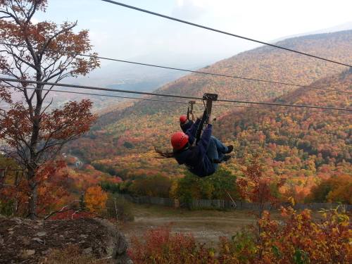 A person zip-lining over a colorful autumn landscape with mountains and trees in vibrant fall colors.