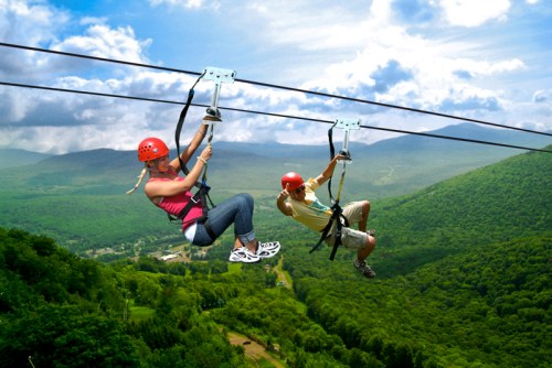 Two people ziplining over a lush green landscape with mountains in the background, wearing helmets and harnesses.