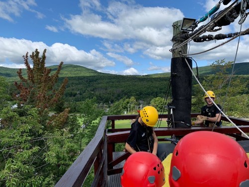 Two people in yellow helmets prepare for a zip line ride, surrounded by green mountains and a blue sky.