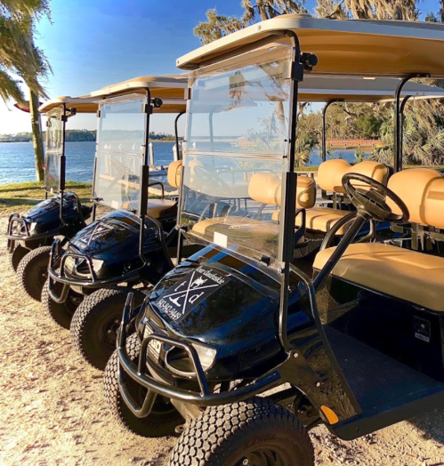 Four golf carts lined up by a lake, featuring tan seats and clear windshields, with trees in the background.