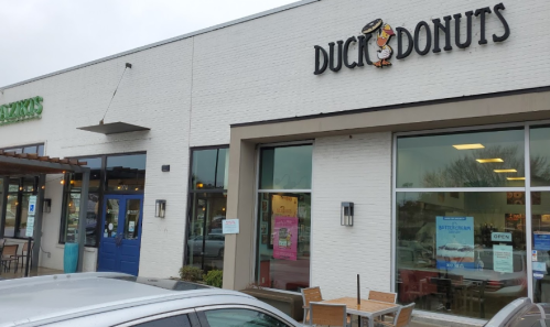 Exterior of Duck Donuts shop with a sign, large windows, and a gray car parked in front. Overcast sky.
