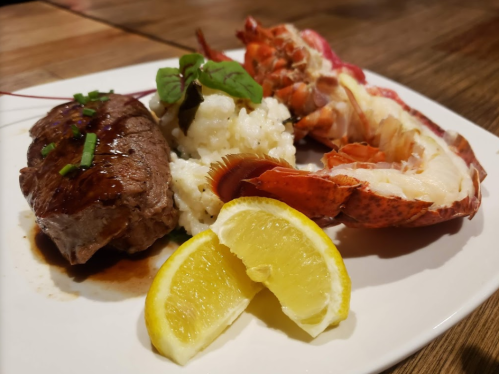 A plate featuring grilled steak, lobster tail, creamy mashed potatoes, and lemon wedges, garnished with herbs.