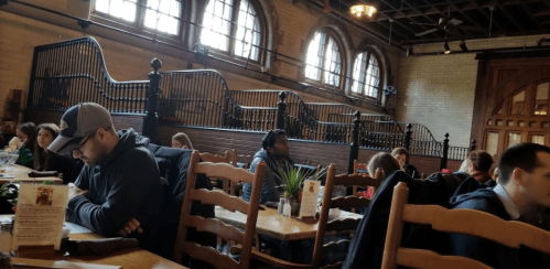 A busy restaurant interior with wooden tables, patrons seated, and large windows letting in natural light.