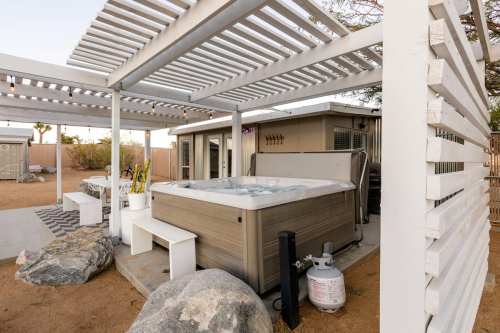 A modern hot tub under a pergola, surrounded by rocks and a patio area with outdoor furniture.