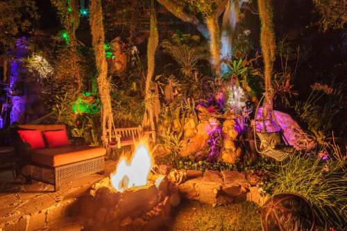 A cozy outdoor space with a fire pit, surrounded by lush greenery and colorful lights, featuring hanging chairs and a sofa.
