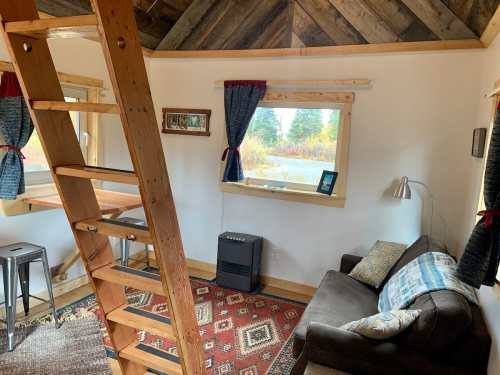 Cozy interior of a small cabin featuring a ladder, sofa, table, and large windows with scenic views.