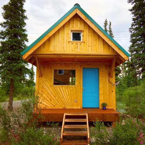 A bright yellow wooden cabin with a teal roof and door, surrounded by greenery and a gravel path.