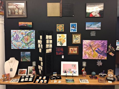 A display of various artworks and crafts on a black wall, featuring paintings, jewelry, and decorative items.